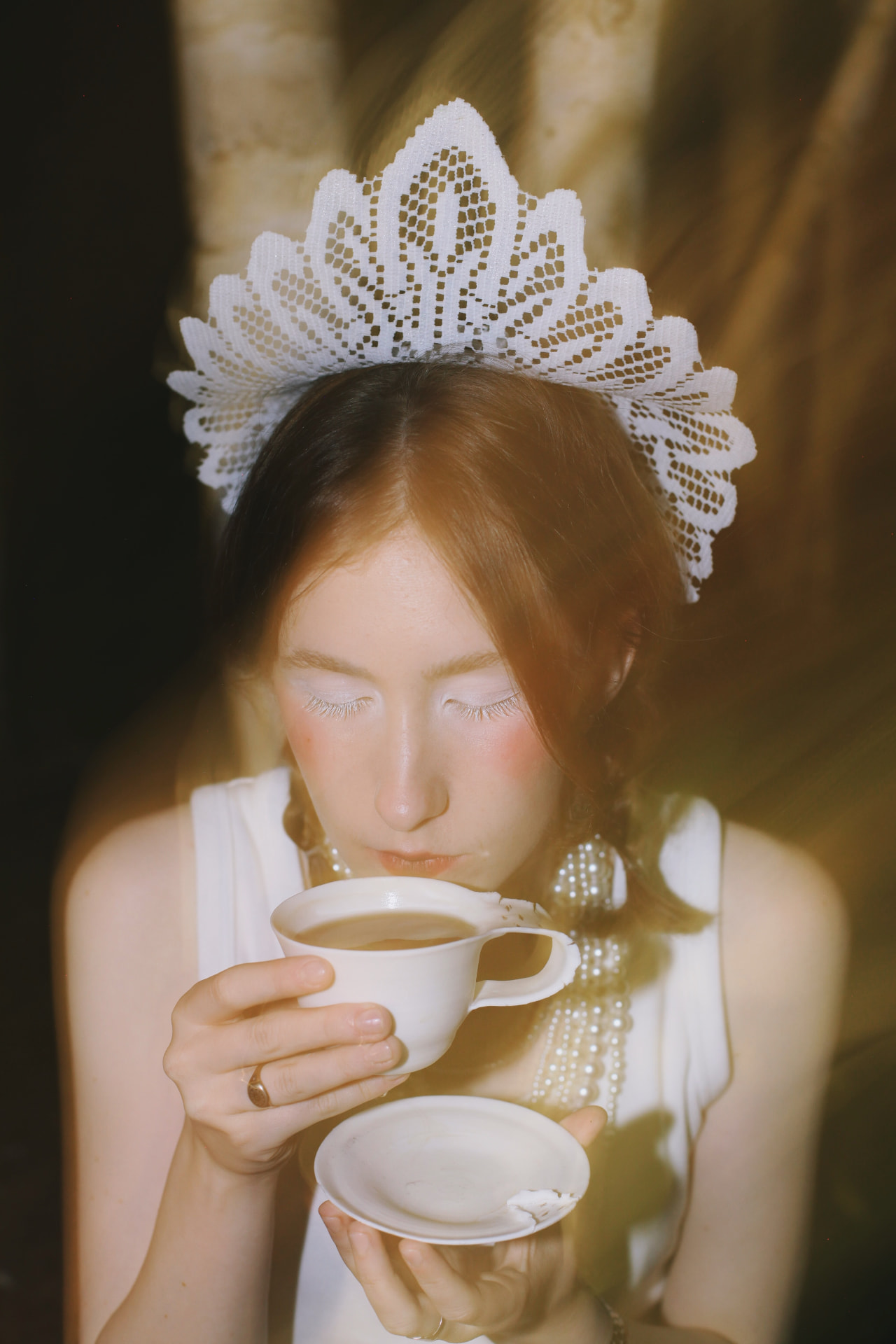 Film portrait — a woman in a white lace halo crown and pearl necklaces drinking from a porcelain teacup