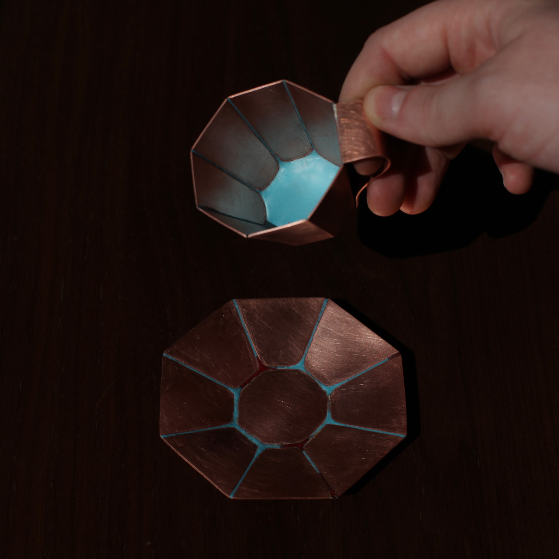 Hand holding a faceted copper cup with turquoise enamel inside, above its matching octagonal saucer on a dark wooden table