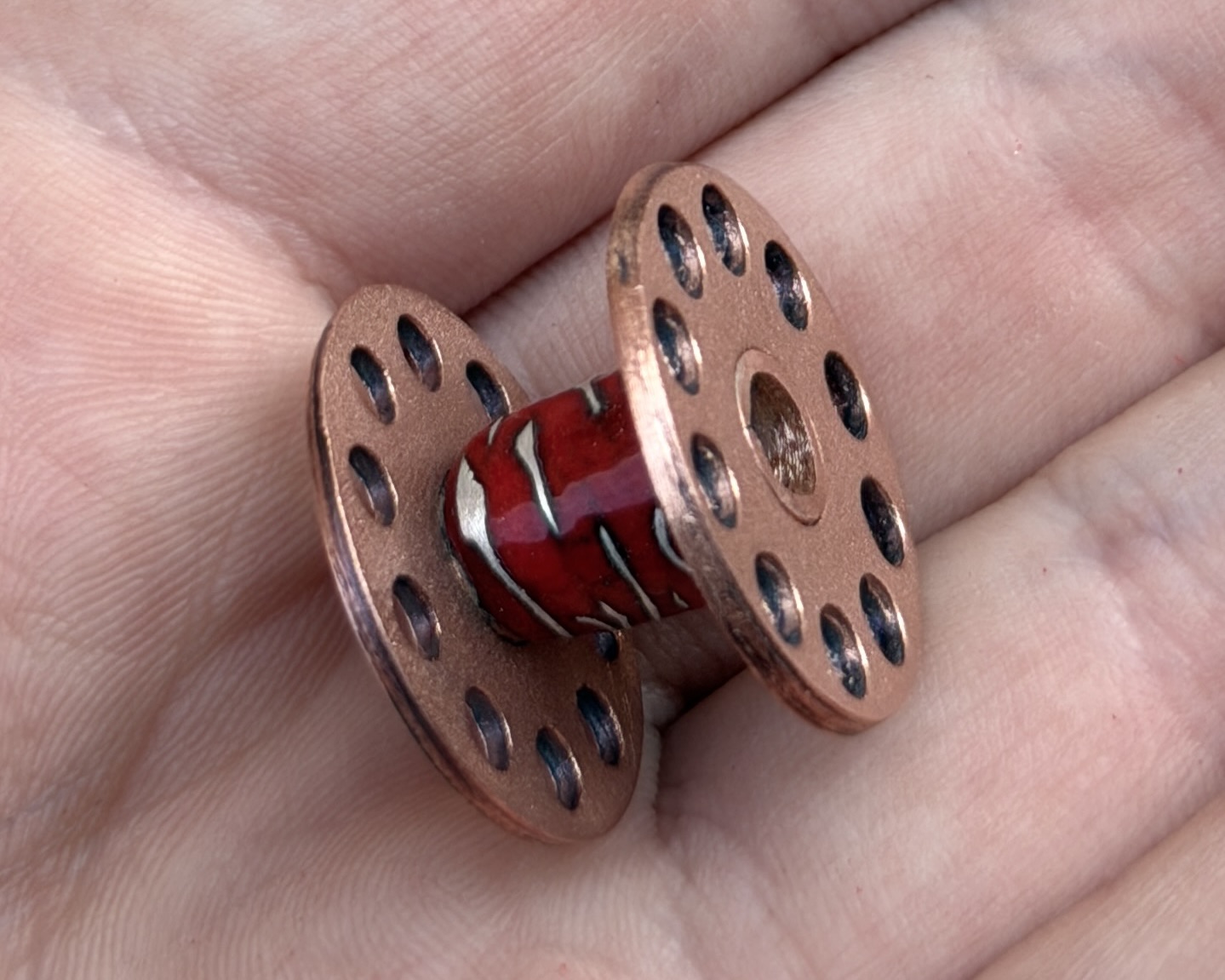 Copper bobbin with a deep red enamel core streaked with silver wire, resting in an open palm