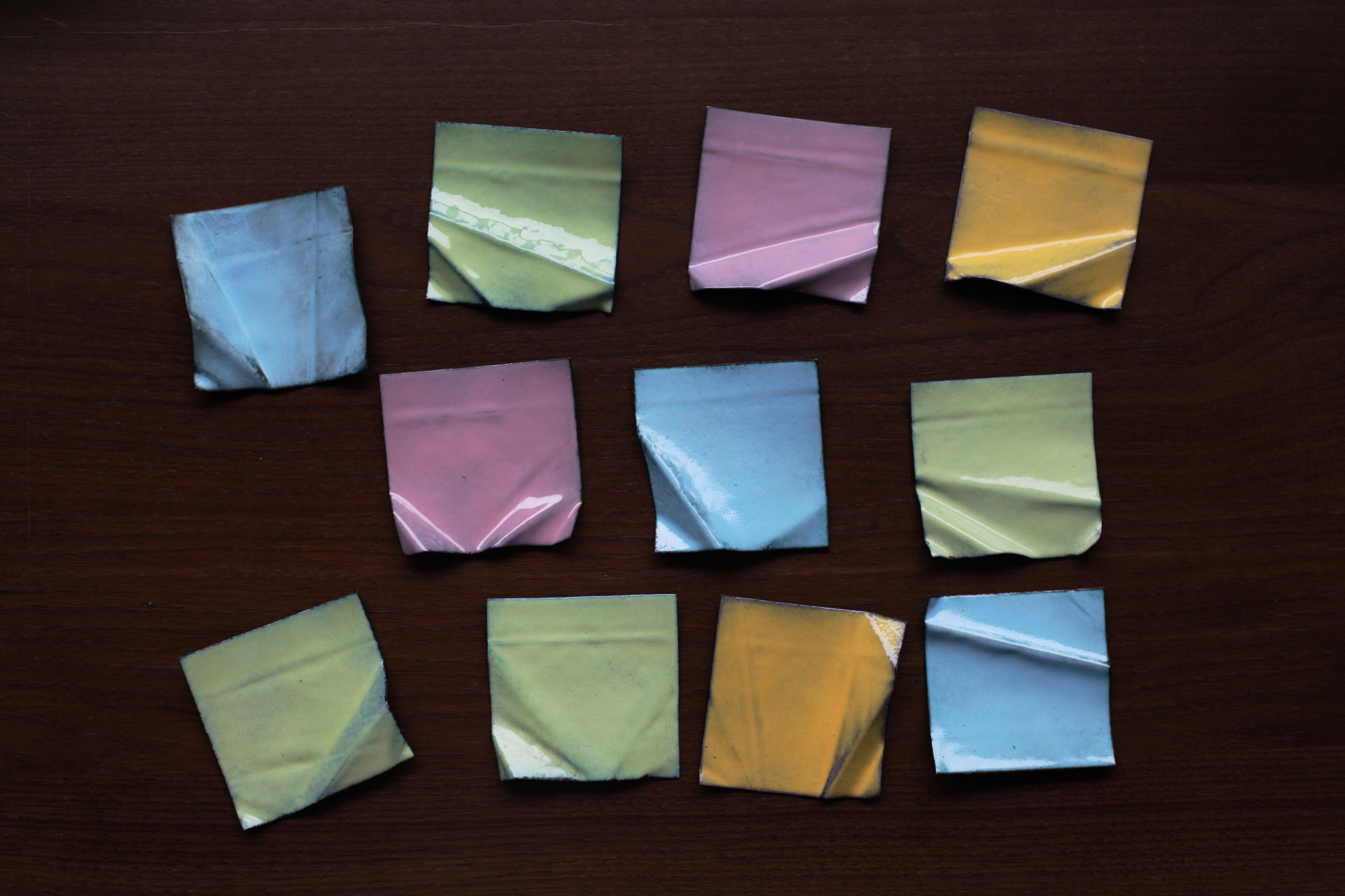 Twelve small pastel enameled copper squares in blue, pink, yellow and green arranged in rows on a dark wooden table