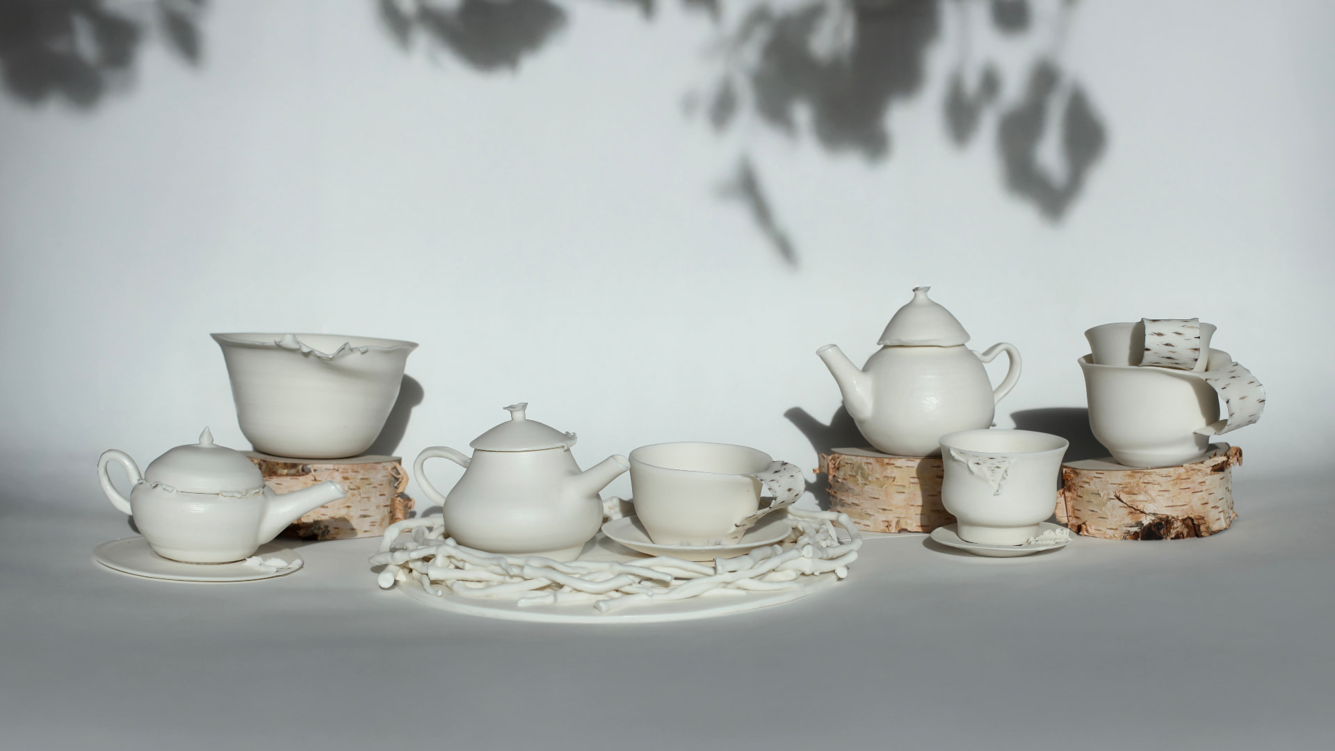 Fine porcelain tea set — teapots, cups and a bowl arranged on birch logs against a white wall