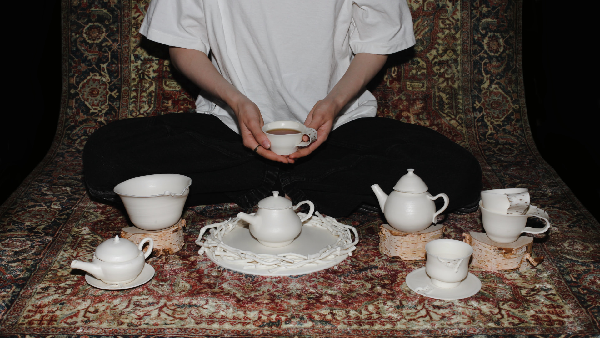 Porcelain tea set laid out on an ornate rug, a person seated cross-legged holding a cup of tea