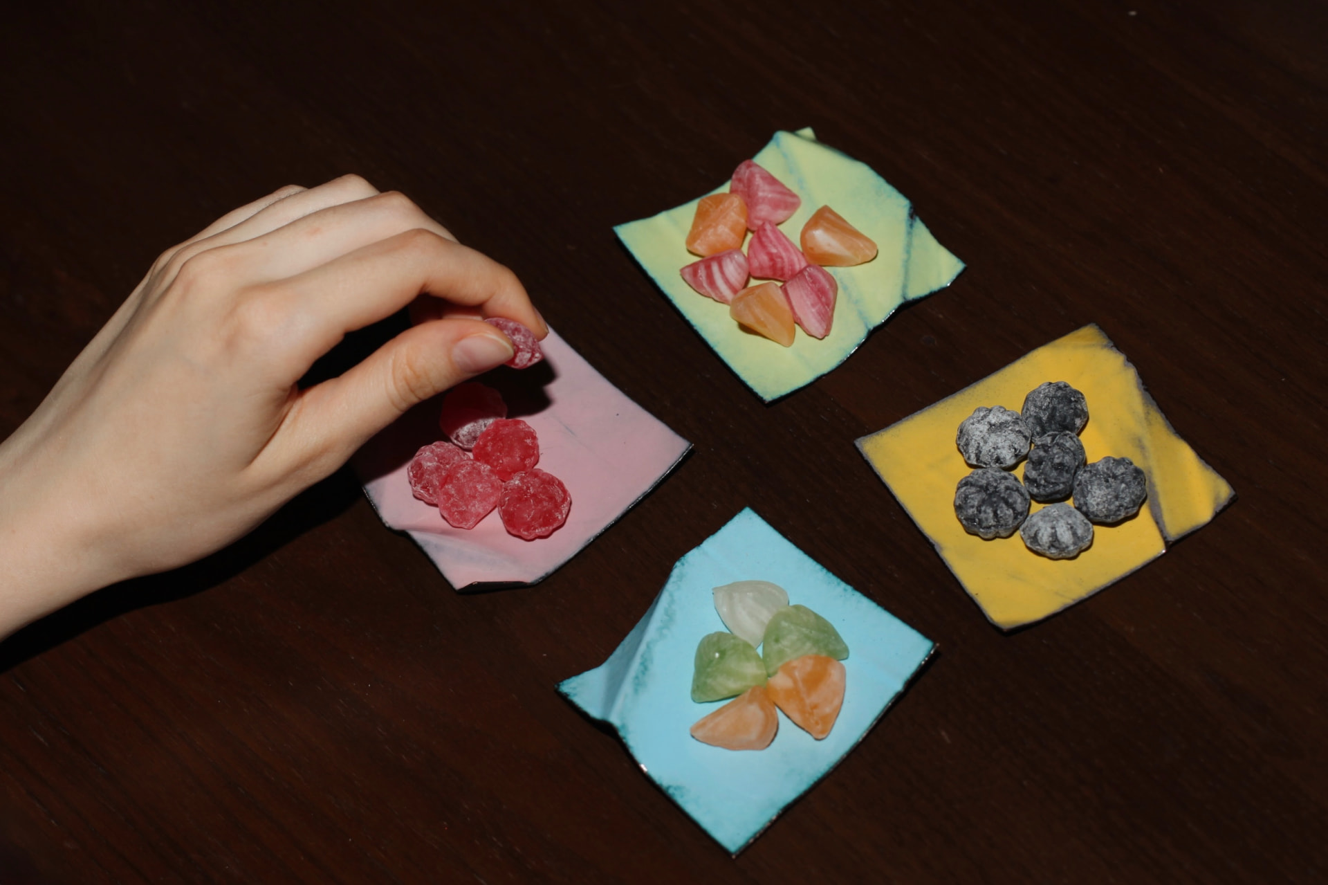 Hand picking a red candy from a small pink enameled copper square, three more colorful squares with sweets on a dark wooden table