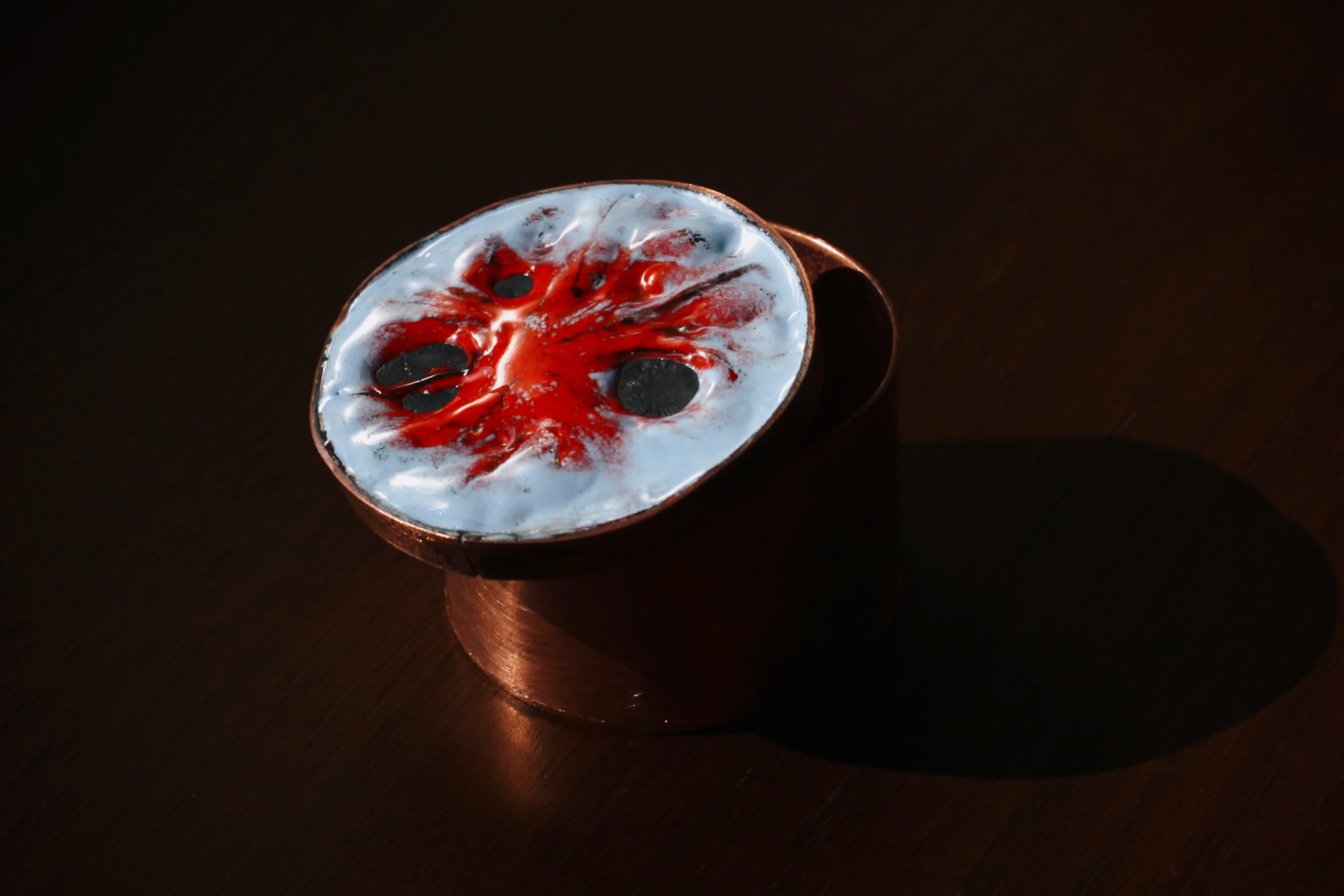 Cylindrical copper box with its enameled lid resting half open — a crimson burst spreading across pale blue-white enamel, on a dark wooden surface